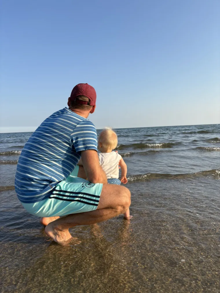 Wir am Wasser: in der Hocke am Ufer, mit unserem Kleinsten auf dem Arm.
